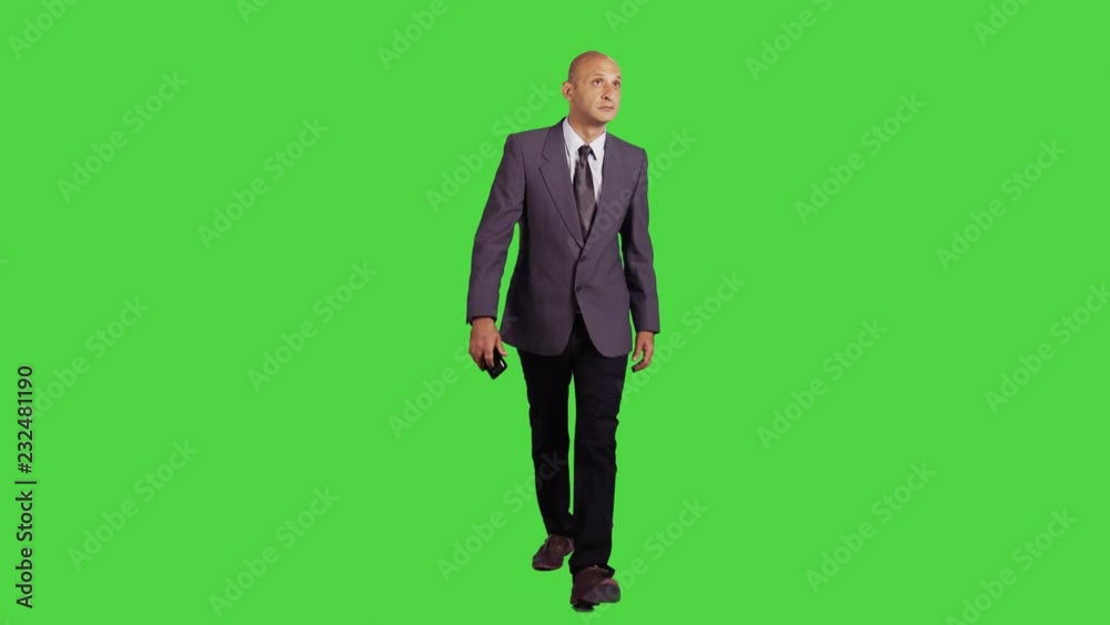Businessman walking towards camera relaxed and holding a phone in a full frontal shot over a green screen, wearing a suit. Businessman walking towards camera relaxed and holding a phone in a full frontal shot over a green screen, wearing a suit.