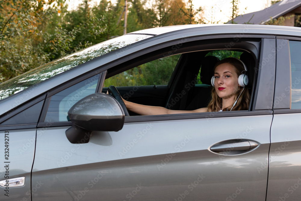 A young, beautiful woman with long hair and headphones sits at the wheel of the car.
