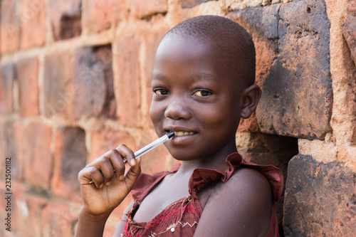 Slika na platnu An 11-year old Ugandan girl smiling, holding a pen against her mouth and leaning
