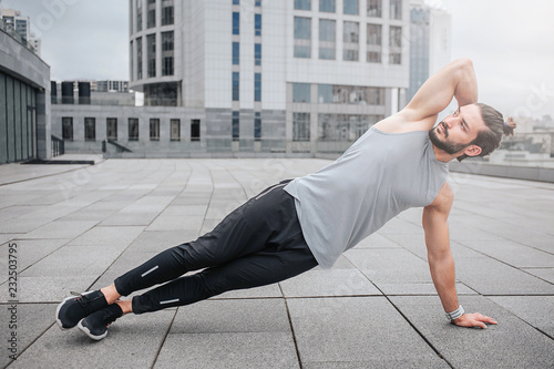 Picture of well-bilt young man stretching his body by doing exercises. He holds one hand behind head. Guy looks forward. He is concentated. Young man exercises alone. photo