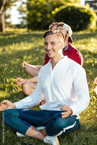 Diverse yoga class of cheerful young friends in summer casual wear sitting in lotus pose on green lown in the public park, front view with focus on leader photo