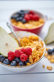 Golden cornflakes with fresh fruits of raspberries, blueberries and pear in two ceramic bowls. Prepared for healthy breakfast. Placed on white wooden table.
