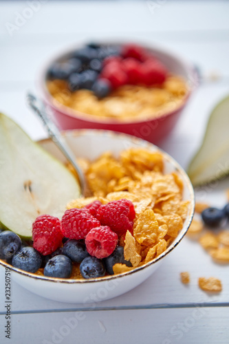 Golden cornflakes with fresh fruits of raspberries, blueberries and pear in two ceramic bowls. Prepared for healthy breakfast. Placed on white wooden table.