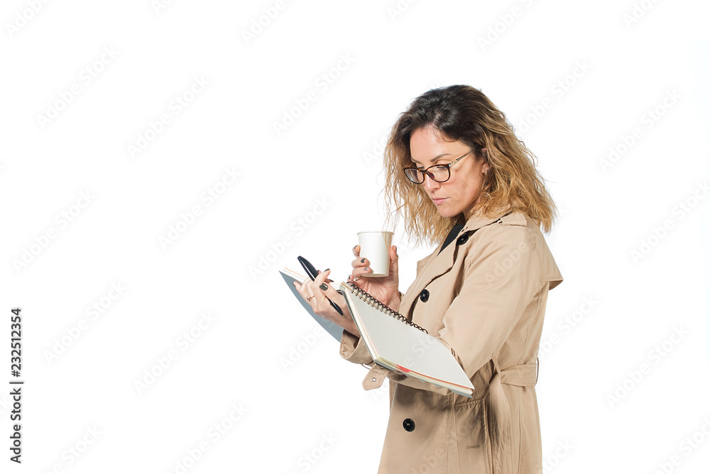 beautiful business woman in glasses and autumn coat isolated on white background. holding a cup of coffee, a notebook, a phone. makes an entry in notebook. checks report. happy face. serious face beautiful business woman in glasses and autumn coat isolated on white background. holding a cup of coffee, a notebook, a phone. makes an entry in notebook. checks report. happy face. serious face