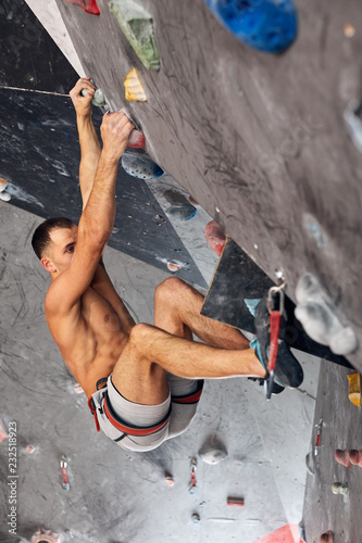 Muscular shitrless man climbing wall with belay device at an indoor wall climbing centre. Rock climber practicing climbing at an indoor climbing gym. photo