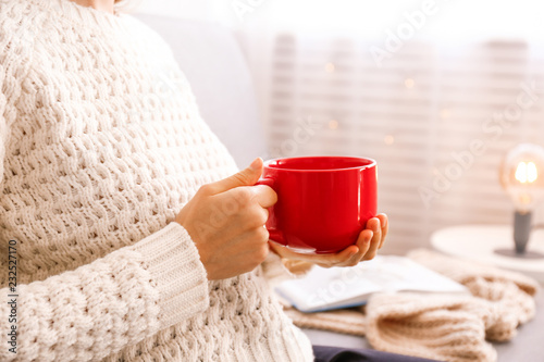 Close up young woman's hands holding big cup of hot beverage. Female wearing fashionable oversized white knitted sweater, sitting home with mug of coffee. Background, copy space.