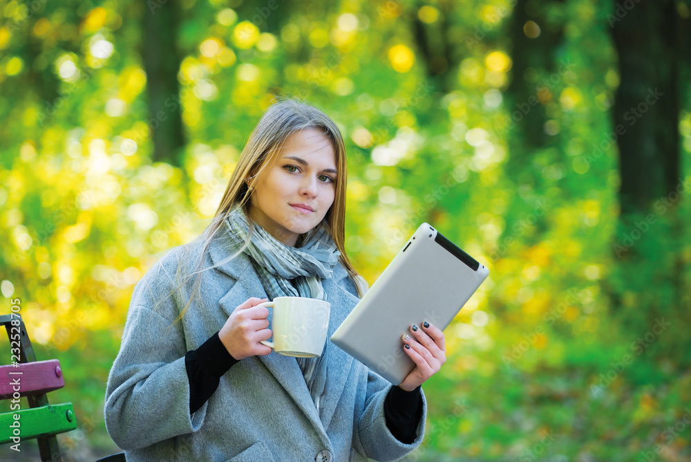 Young blonde girl with umbrella sitting on a park bench in the autumn season. girl in a gray coat drinks coffee, looks into a tablet, talks on the phone, reads a book, writes in a notebook