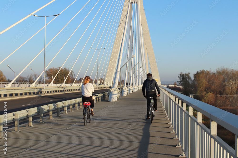 Rzeszow, Poland - 9 9 2018: A guy with a girl riding bicycles on a suspension road bridge across the Wislok River on a sunny, clear day. Metal construction technological structure in Europe.
