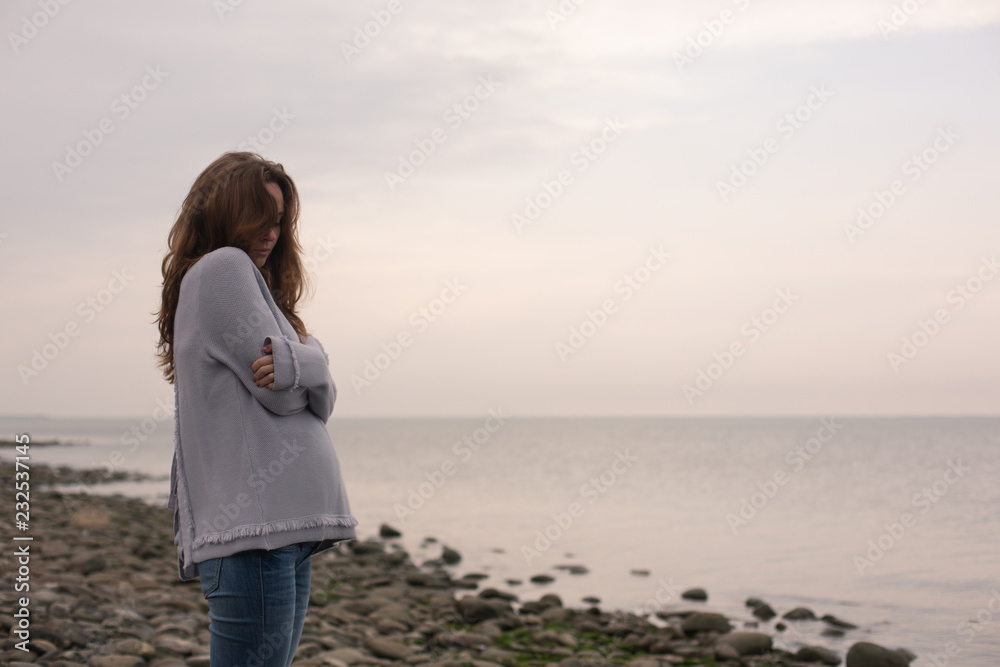 Young Sad Pregnant Woman in a Knitted Sweater Standing on a Deserted Beach. Sea on a Background. Young Sad Pregnant Woman in a Knitted Sweater Standing on a Deserted Beach. Sea on a Background.