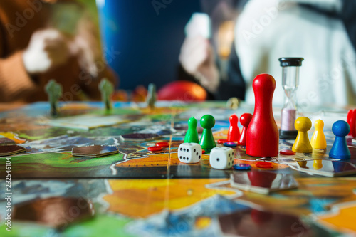 Board game concept- board game field, many figures, meeple, dice, coins and sandglass. Two people play holding cards on blurred background photo