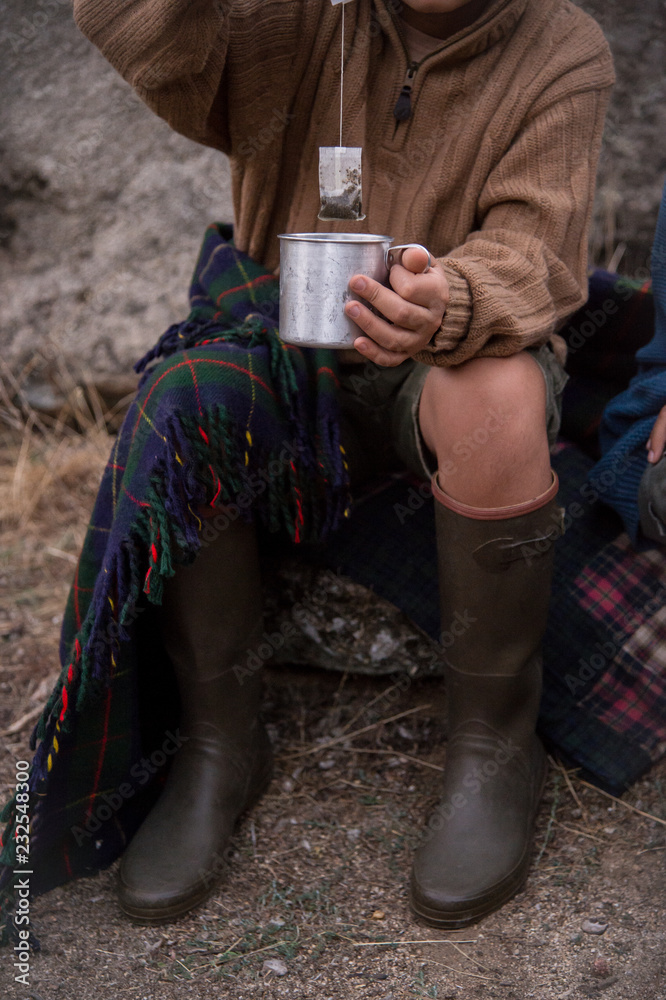 Boy holding a tea bag over a metal cup covered by a plaid blanket and wearing a wool sweater and boots. Boy holding a tea bag over a metal cup covered by a plaid blanket and wearing a wool sweater and boots.