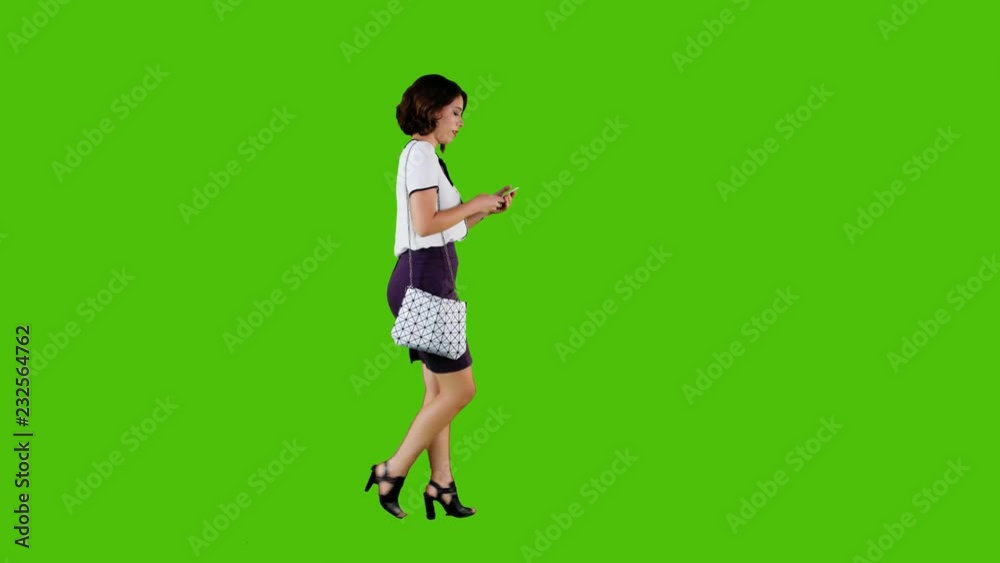 Stylish woman using an app cheerfully while walking over a green screen in in a full side shot. Stylish woman using an app cheerfully while walking over a green screen in in a full side shot.