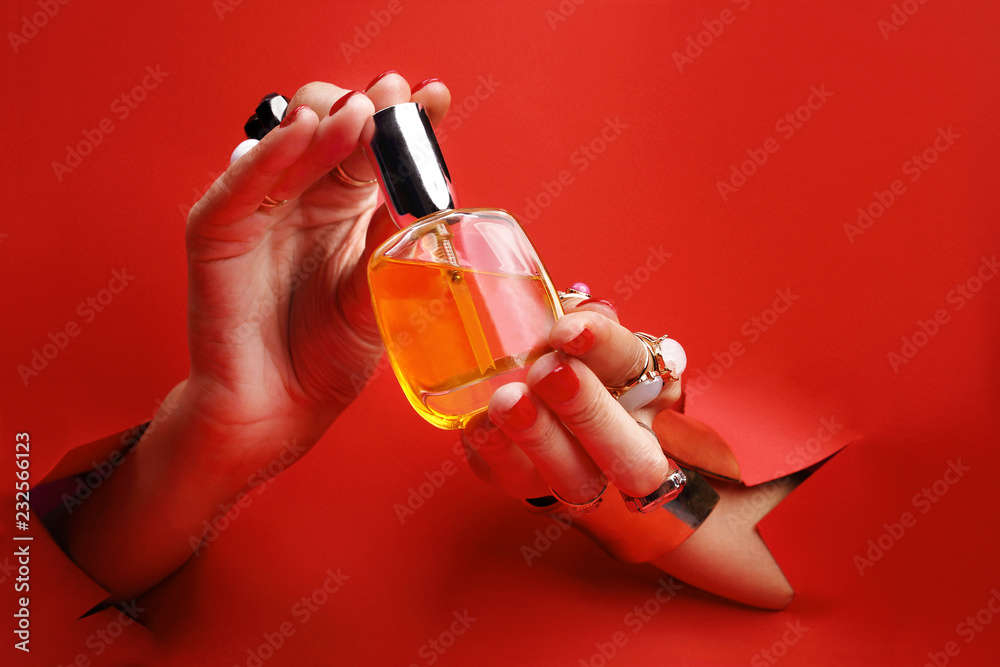 Eleganckie kosmetyki w kobiecych dłoniach.  Kobiece dłonie z  czerwonymi paznokciami przez otwór w czerwonym tle  trzymają perfumy. - obrazy, fototapety, plakaty 