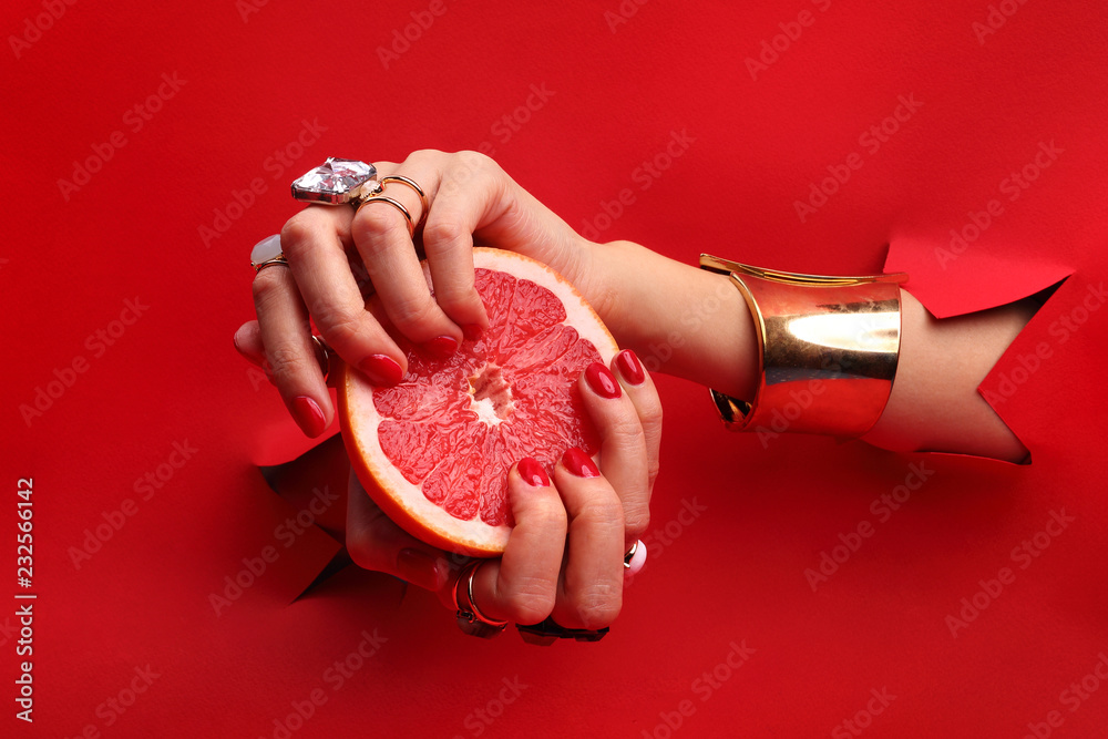 Manicure. Kobiece dłonie z  czerwonymi paznokciami przez otwór w czerwonym tle  trzymają owoc grejpfruta. - obrazy, fototapety, plakaty 