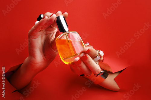 Eleganckie kosmetyki w kobiecych dłoniach. Kobiece dłonie z czerwonymi paznokciami przez otwór w czerwonym tle trzymają perfumy.