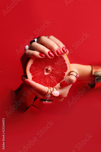 Kobiece eleganckie dłonie. Kobiece dłonie z czerwonymi paznokciami przez otwór w czerwonym tle trzymają owoc grejpfruta.