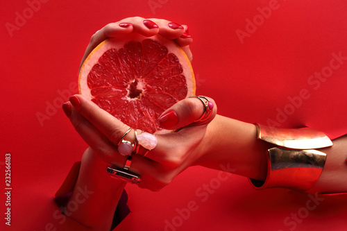 Kobiece eleganckie dłonie. Kobiece dłonie z czerwonymi paznokciami przez otwór w czerwonym tle trzymają owoc grejpfruta.
