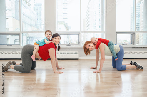 Group of two young women with children doing workout in gym class to loose baby weight. Child-friendly fitness for mothers with kids toddlers. Lifestyle concept of parent activity