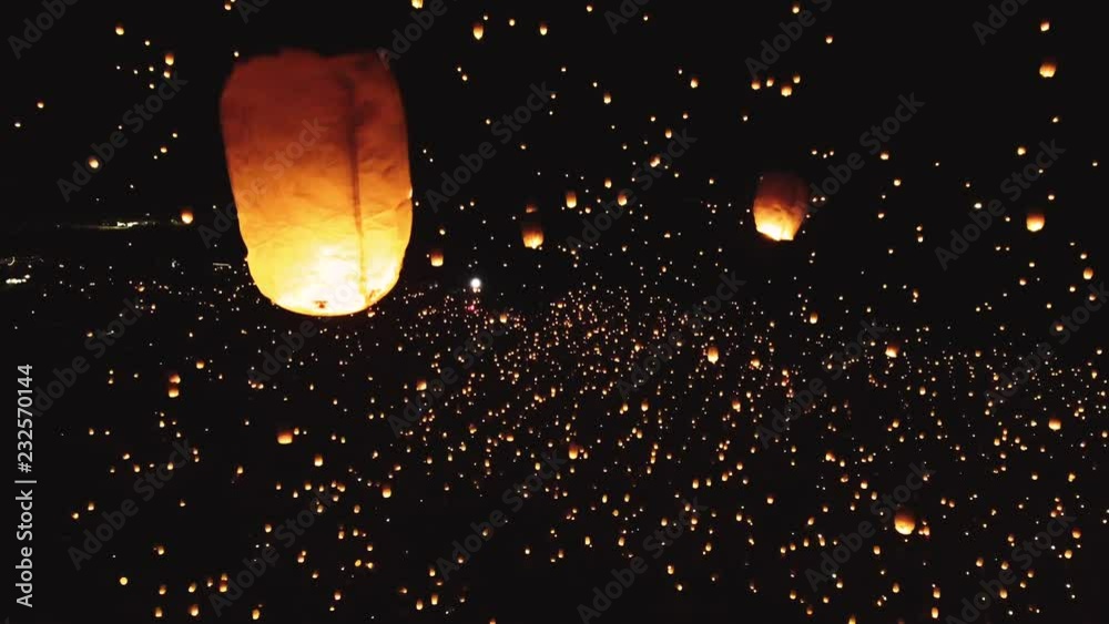 Drone shot of thousands of glowing lanterns flying across screen. Beautiful floating lanterns light the night sky as thousands of people gather throw fire lanterns in the sky.