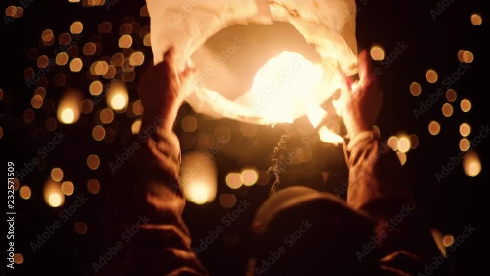 A person grabbing and letting go of a lantern sending it off into the night sky. A lantern lit by fire flys slowly off into the air. Close up up a persons hands holding a lantern.