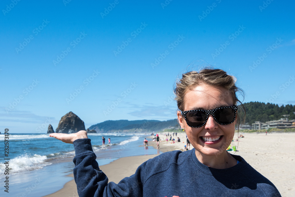 Woman wearing sunglasses pretends to hold up a seastack rock in Cannon Beach, Oregon on the beach on a sunny day. Forced perspective view