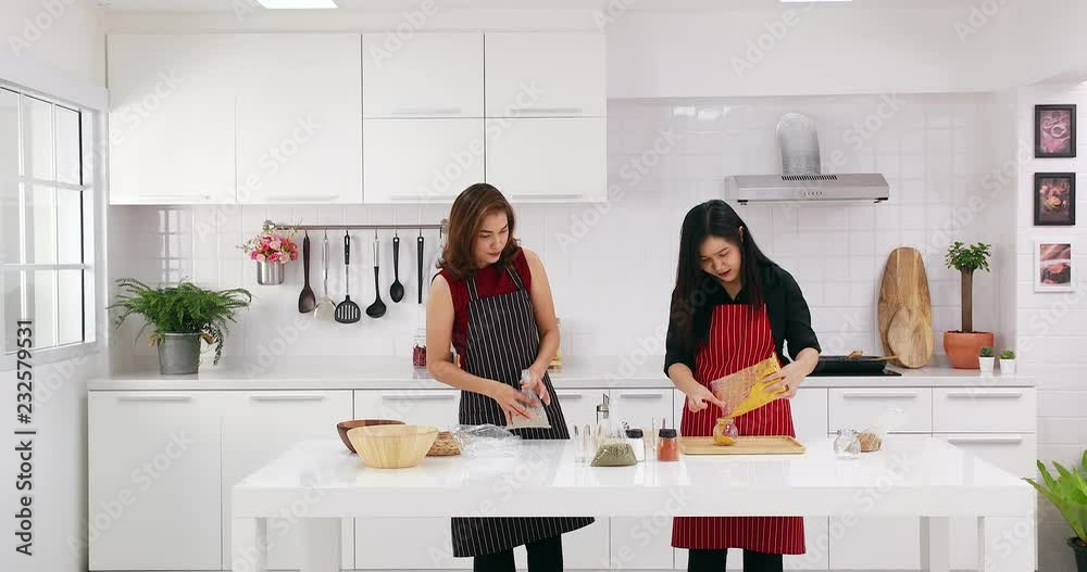 Good looking Asian women in kitchen, red apron woman pour some ingredients into jar, black apron woman bind some plastic bag and take it out of kitchen, concept for Asian women in kitchen. Good looking Asian women in kitchen, red apron woman pour some ingredients into jar, black apron woman bind some plastic bag and take it out of kitchen, concept for Asian women in kitchen.