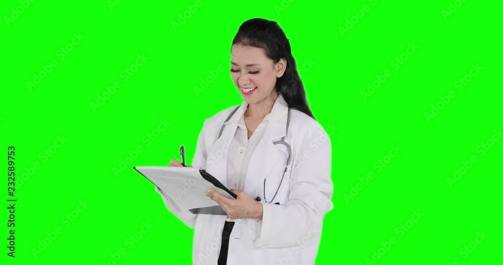Beautiful young doctor standing in the studio while writing on a clipboard with green screen background. Shot in 4k resolution Beautiful young doctor standing in the studio while writing on a clipboard with green screen background. Shot in 4k resolution