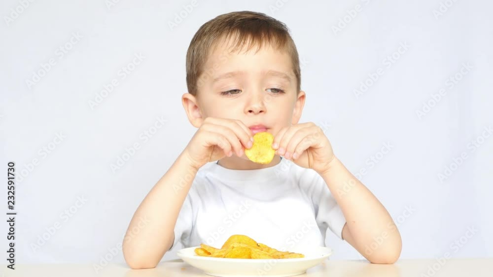 The child with pleasure and joy, eating potato chips, sitting at the table on a white background. Boy loves crispy potato chips. The child with pleasure and joy, eating potato chips, sitting at the table on a white background. Boy loves crispy potato chips.