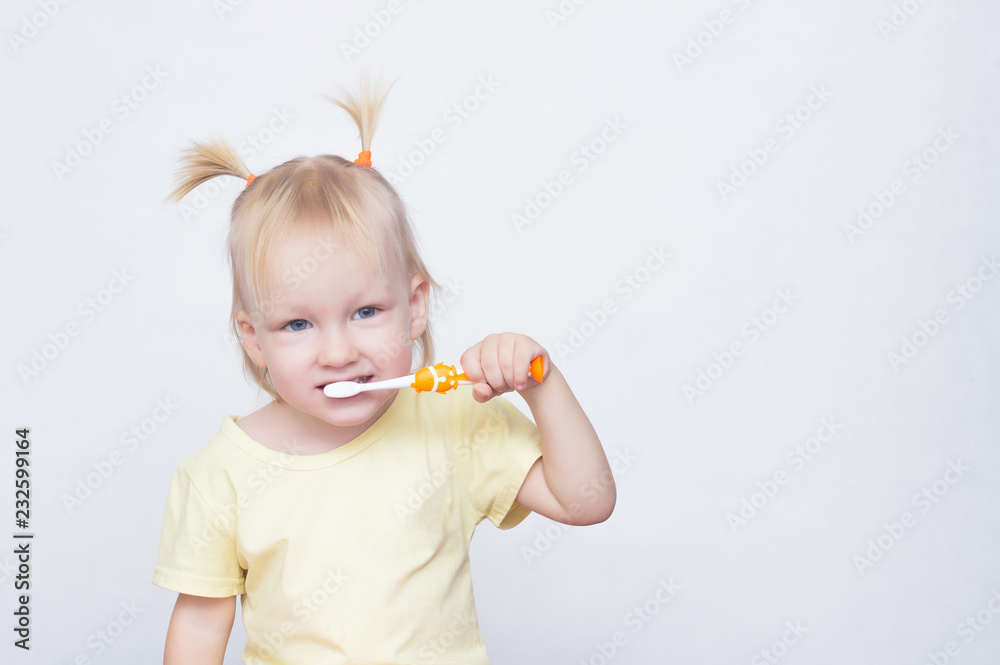 Little blue-eyed girl blonde with pigtails on her head brushing teeth with a toothbrush and looking at the camera, white background, copy space, caucasian Little blue-eyed girl blonde with pigtails on her head brushing teeth with a toothbrush and looking at the camera, white background, copy space, caucasian