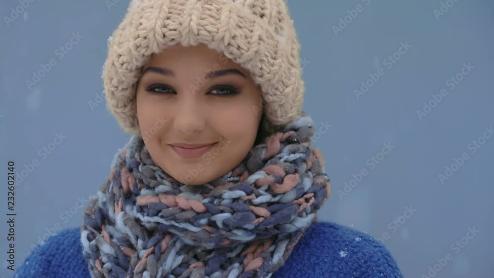 Happy women facing camera during winter holidays outside. Snow falling in slow motion 4K. Young female enjoying winter day outdoors. Woman in cap, coat, scarf standing against blue copy space Happy women facing camera during winter holidays outside. Snow falling in slow motion 4K. Young female enjoying winter day outdoors. Woman in cap, coat, scarf standing against blue copy space