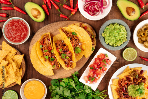A photo of Mexican food, including tacos, guacamole, pico de gallo, nachos and others, shot from the top with ingredients on a dark rustic wooden background