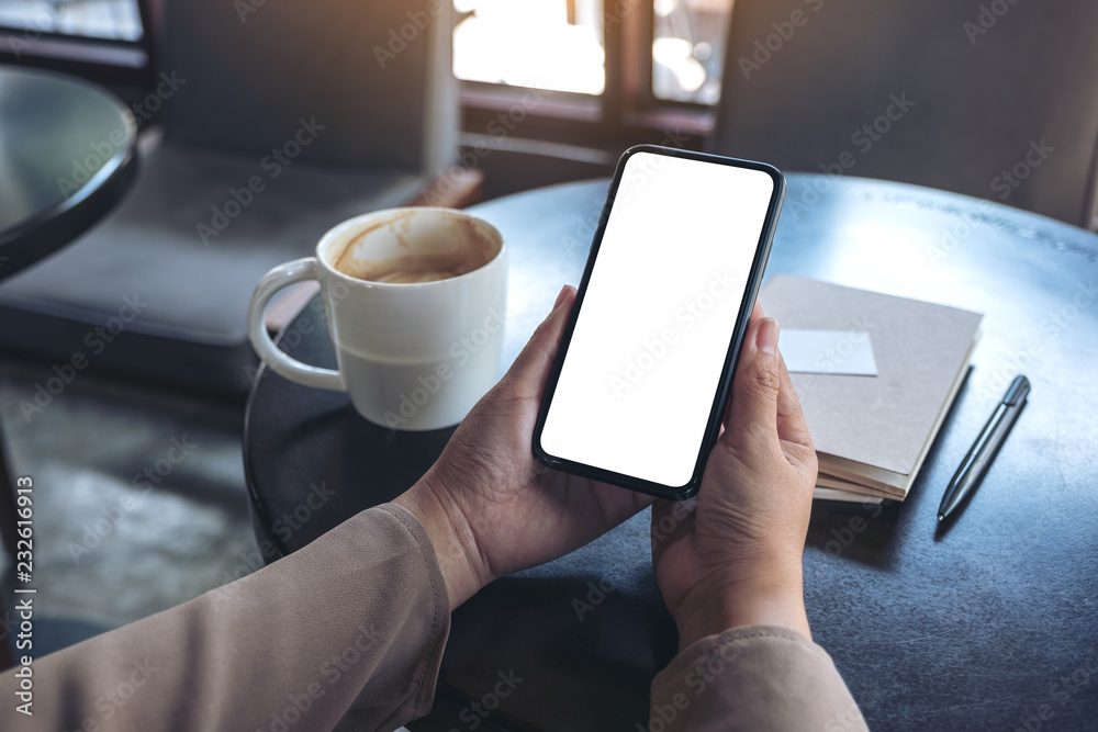 Mockup image of woman's hands holding and using a black mobile phone with blank screen for watching with notebook and coffee cup on wooden table Mockup image of woman's hands holding and using a black mobile phone with blank screen for watching with notebook and coffee cup on wooden table