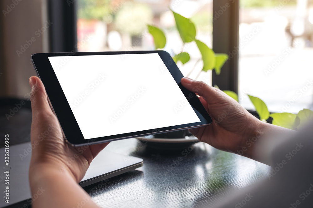 Mockup image of hands holding black tablet pc with blank white desktop screen with green leaves on wooden table Mockup image of hands holding black tablet pc with blank white desktop screen with green leaves on wooden table