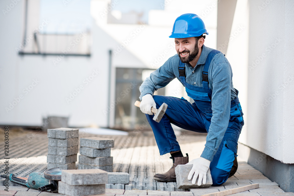 Builder in uniform laying paving tiles on the construction site with white houses on the background Builder in uniform laying paving tiles on the construction site with white houses on the background