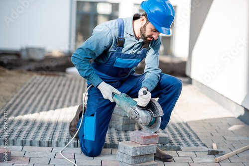 Builder in uniform cutting paving tiles with electric cutter on the construction site with white houses on the background