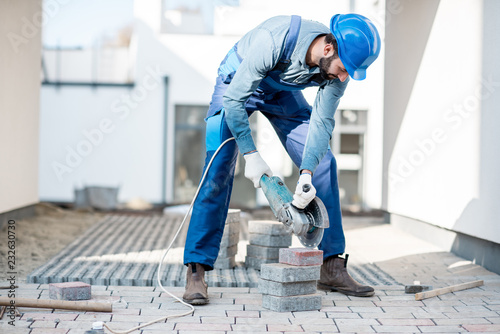 Builder in uniform cutting paving tiles with electric cutter on the construction site with white houses on the background photo