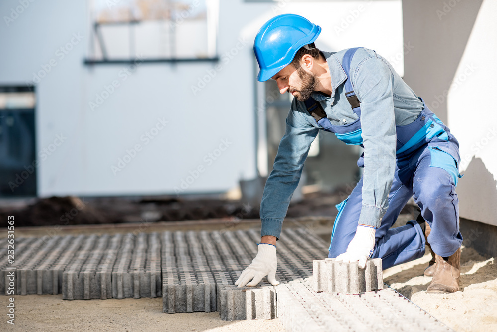 Builder in uniform mounting paving tiles on the construction site with white houses on the background