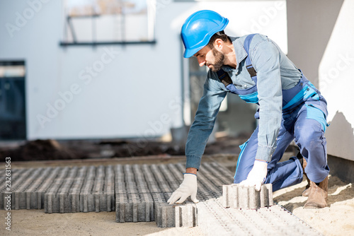Builder in uniform mounting paving tiles on the construction site with white houses on the background