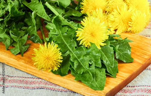 Fresh dandelion leaves with flowers on cutting board on table with linen textile, closeup, copy space. Healthy natural organic food concept photo