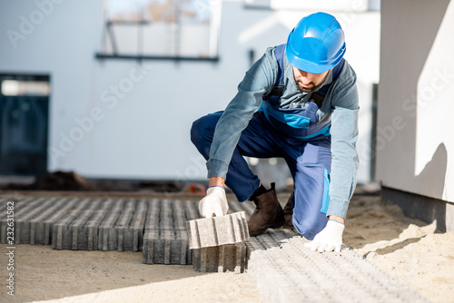 Builder in uniform mounting paving tiles on the construction site with white houses on the background