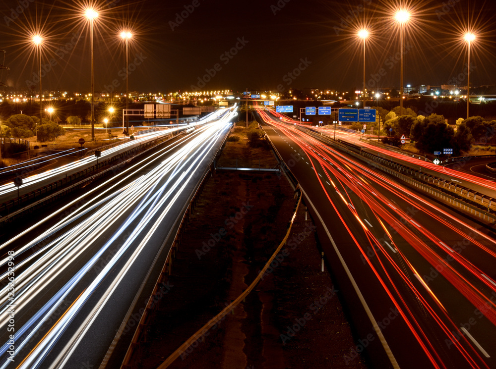 DIFFERENT WHITE, BLUE, YELLOW AND RED TRAFFIC LIGHTS OF MANY VEHICLES IN A SPANISH MOTORWAY WITH THE LIGHT OF A CITY AT THE END OF THE HORIZON