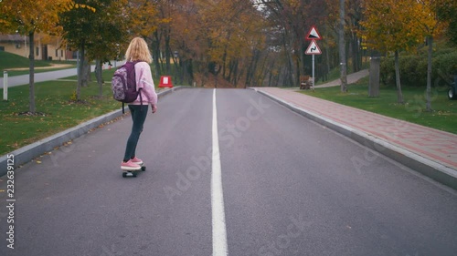 Young pretty beautiful blond hipster woman in pink windstopper having fun riding skateboard longboard downhill on beautiful road in slow motion photo