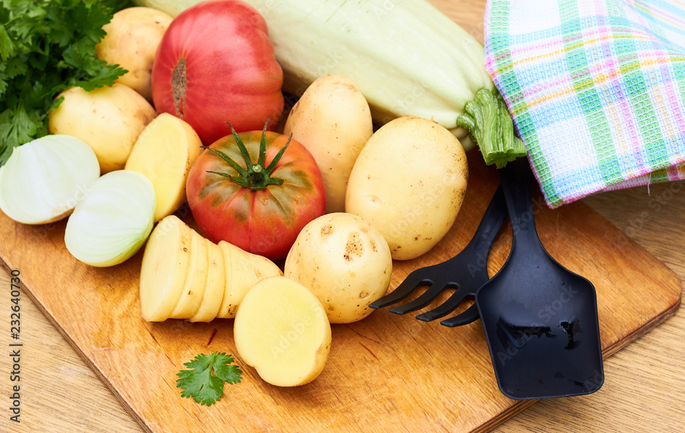 Fresh garden vegetables and herbs: tomatoes, potatoes, onion, parsley, cilantro, zucchini assorted on cutting board on  wooden table with kitchen stuff nearby: napkin, fork and ladle