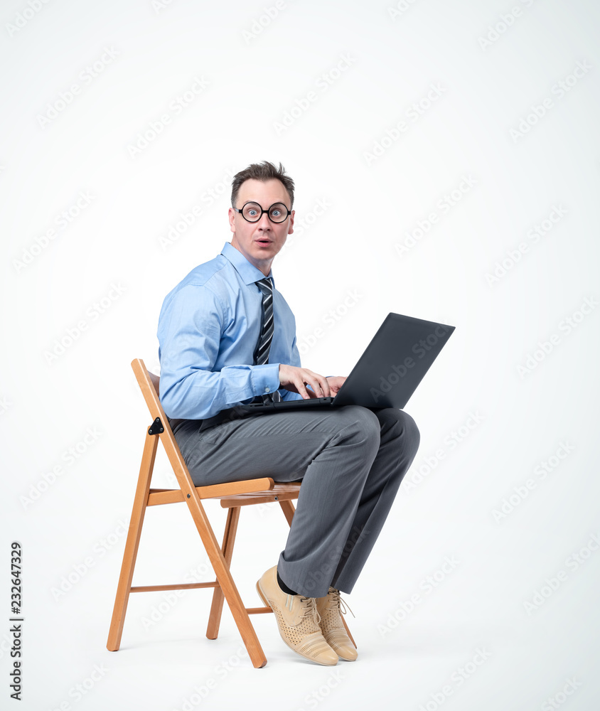 Funny male programmer in glasses is sitting on a chair with a laptop, on background. 