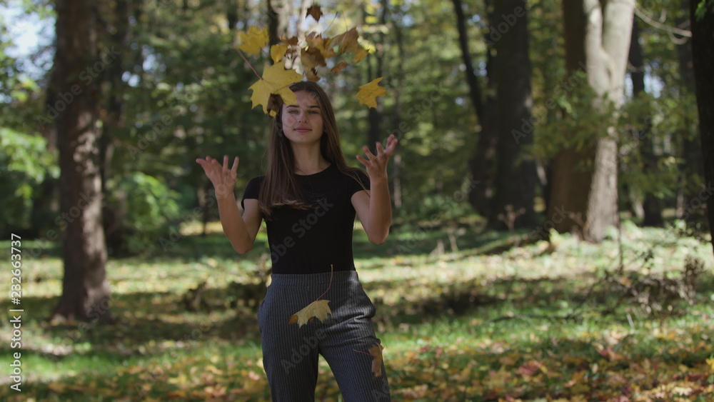 girl is covering her face and waving a maple tree branch with yellow leaves in autumn park. Slow motion