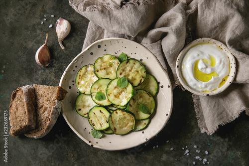 Grilled zucchini salad with yogurt dip and rye sliced bread in spotted ceramic plates on linen cloth over old dark metal texture background. Vegetarian food. Flat lay, space
