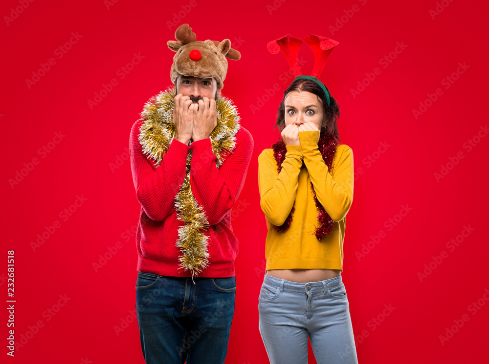 Couple dressed up for the christmas holidays is a little bit nervous and scared putting hands to mouth on isolated red background Couple dressed up for the christmas holidays is a little bit nervous and scared putting hands to mouth on isolated red background