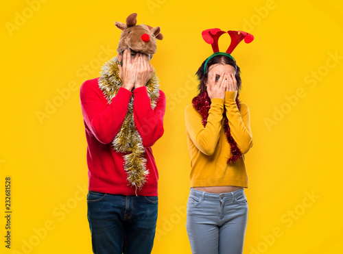 Couple dressed up for the christmas holidays covering eyes by hands and looking through the fingers on yellow background