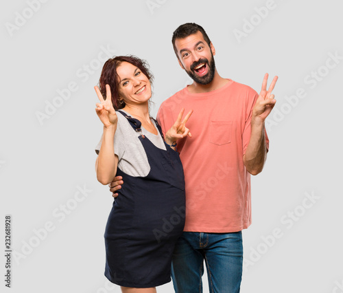 Couple with pregnant woman smiling and showing victory sign with both hands on isolated grey background