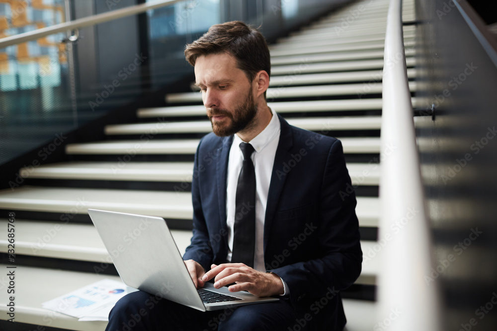 Young mobile businessman sitting on stairs and looking at data on laptop display while networking Young mobile businessman sitting on stairs and looking at data on laptop display while networking